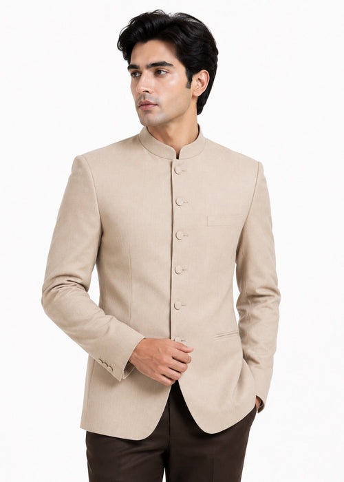 A man wearing a light beige mandarin collar Nehru jacket with buttoned front closure, paired with dark brown trousers, photographed against a plain white background.